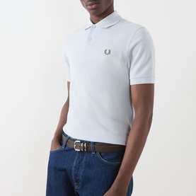 Model wearing Fred Perry Twin Tipped Polo Shirt in Dusty Blue with Laurel Wreath Green logo, styled with blue jeans and brown belt.