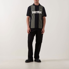 Model wearing Fred Perry Meyba Cut & Sew Polo Shirt in black with khaki stripe panels and graphic print, styled with black trousers and loafers