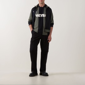 Model wearing Fred Perry Meyba Cut & Sew Polo Shirt in black with graphic print, styled with khaki zip jacket and black trousers