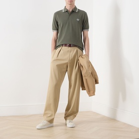Model wearing Fred Perry Twin Tipped Logo Polo Shirt in Laurel Wreath Green with ecru tipping, styled with beige wide-leg trousers and white trainers.