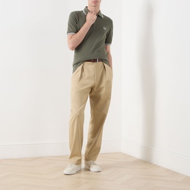 Model wearing Fred Perry Twin Tipped Logo Polo Shirt in Laurel Wreath Green with ecru tipping, styled with beige pleated trousers and white trainers.