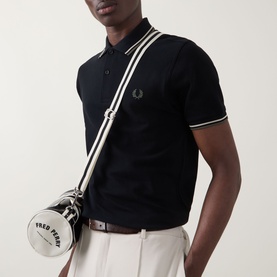 Model wearing Fred Perry Laurel Logo Twin Tipped Polo Shirt in Black with white tipped collar, carrying cylindrical Fred Perry crossbody bag, styled