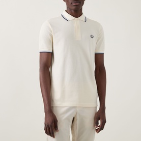 Model wearing cream polo shirt with navy twin tipping and embroidered laurel wreath logo, styled with matching cream trousers