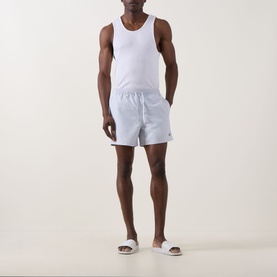 Model wearing Fred Perry Laurel Logo Swim Shorts in Dusky Blue with white vest top and white slider sandals.