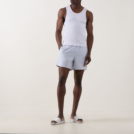 Model wearing Fred Perry Laurel Logo Swim Shorts in Dusky Blue with white vest top and white slider sandals.