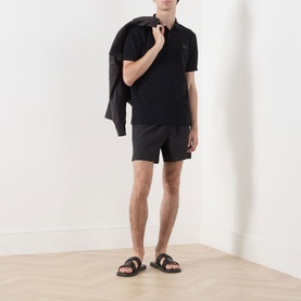 Model wearing Fred Perry Laurel Wreath Swim Shorts in Black with a black polo shirt and dark slides, holding a jacket over one shoulder.