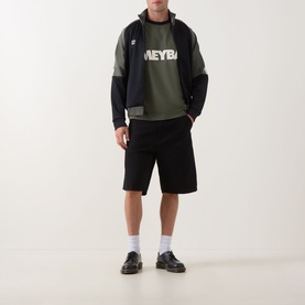 Model wearing Fred Perry Meyba Taped Track Jacket in black, two-tone zip-through style, styled with khaki graphic sweatshirt and black shorts.