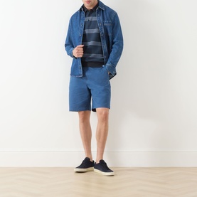 Model wearing Scotch & Soda Ensign Blue chino shorts, styled with a denim shirt, striped polo, and navy canvas shoes.