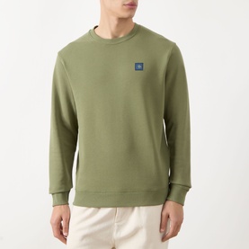 Model wearing Scotch & Soda Seasonal Essentials Logo Badge Sweatshirt in Oil Green, crew neck with logo badge, paired with cream trousers.