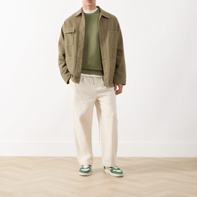 Model wearing Scotch & Soda Seasonal Essentials Logo Badge Sweatshirt in Oil Green, styled with khaki jacket, cream trousers and green trainers.