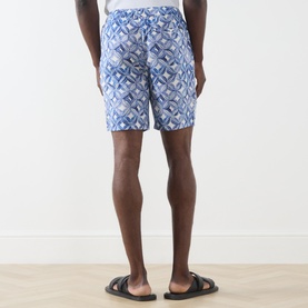 Model wearing Scotch & Soda Tile Flower Printed Bermuda Shorts in blue geometric print, styled with black leather sandals, rear view.