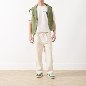 Model wearing Scotch & Soda Artwork Logo T-Shirt in Eggnog, styled with a green vest, cream trousers, and green trainers.