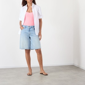 Model wearing light blue denim longline shorts, styled with a pink vest top, white linen shirt, and tan flat sandals.