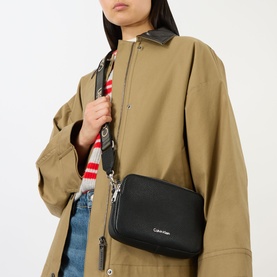 Model carrying Calvin Klein Logo Camera Crossbody Bag in black with silver-tone hardware, styled with wide webbed crossbody strap over a beige coat.