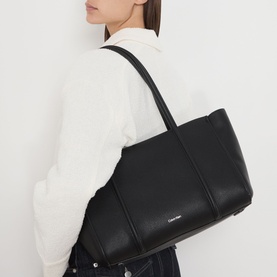 Model carrying Calvin Klein Monogram Emblem Tote Bag in black faux grained finish with silver-tone hardware and shoulder strap, styled with white top