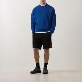 Model wearing Hugo black cotton shorts with red logo patch, styled with blue sweatshirt and black trainers