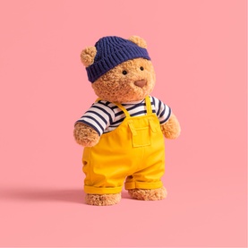 Jellycat small teddy bear plush wearing yellow dungarees, navy striped top and knitted beanie hat, against pink background