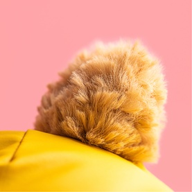 Jellycat plush toy close-up showing fluffy golden-brown pompom detail on yellow fabric, against pink background