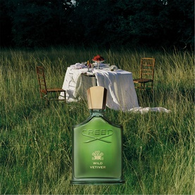 CREED Wild Vetiver Eau De Parfum 100ml, green glass bottle with gold cap, styled in a meadow with an outdoor dining scene behind