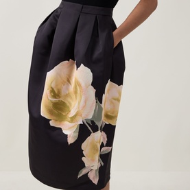 Model wearing Ted Baker Verinie Placement Floral Dress in black, showing pleated midi skirt with large rose placement print and satin finish.