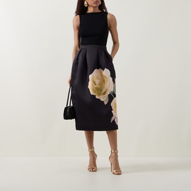 Model wearing Ted Baker Verinie Placement Floral Dress in black, midi length with large floral print skirt, styled with gold heeled sandals and black