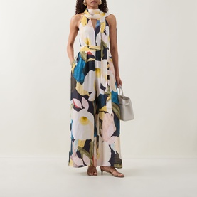 Model wearing Ted Baker Camea Floral Print Jumpsuit in Navy with halter neck, tie detail and wide-leg silhouette, styled with a grey bag and heels.