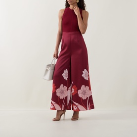 Model wearing Ted Baker Izeny Placement Floral Jumpsuit in dark red, sleeveless, wide-leg with floral print. Styled with nude heels and white bag.