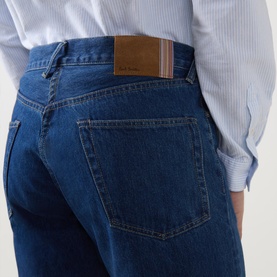 Model wearing Paul Smith blue denim jeans, rear view showing brown leather patch label and five-pocket styling, paired with a striped shirt.