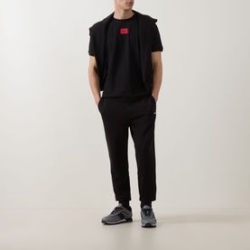 Model wearing Hugo black cotton T-shirt with red logo patch, styled with matching black joggers and grey trainers, layered under a black gilet.