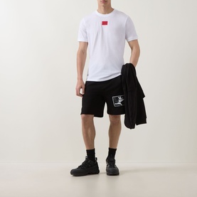 Model wearing Hugo white cotton T-shirt with small red chest logo, styled with black shorts and black chunky trainers, carrying black jacket