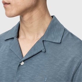 Model wearing All Saints lunar blue short sleeve camp collar shirt, close-up of neckline and embroidered logo detail