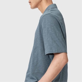 Model wearing All Saints short-sleeve polo shirt in Lunar Blue, textured marl fabric, relaxed fit, side and back view.