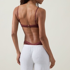 Model wearing Tala Deep Rust bikini top, back view with gold buckle detail, styled with white wide-leg trousers.