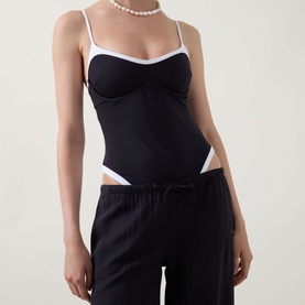 Model wearing Tala Contrast Trim Swimsuit in Shadow Black, one-piece with white contrast trim, V-neckline and slim straps, styled with black trousers.