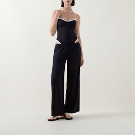 Model wearing Tala Contrast Trim Swimsuit in Shadow Black with white contrast trim and slim straps, styled with wide-leg black trousers and sandals.
