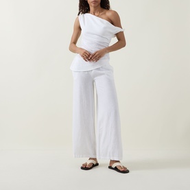 Model wearing Tala ivory one-shoulder linen blend top, styled with matching wide-leg white trousers and flat sandals.