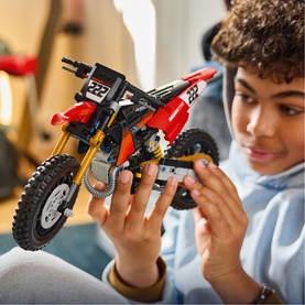 Child assembling Lego Technic dirt bike model in red, black and gold, holding the detailed motocross build with number 222 on the front.