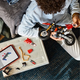 Child assembling Lego Technic motocross bike model, with loose pieces and instruction booklet on tray nearby, overhead view