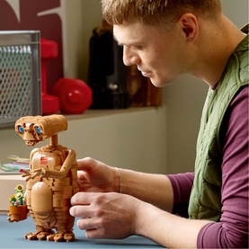 Man assembling Lego Ideas wooden-style robot figure at a desk, with small character builds visible nearby