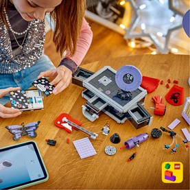 Child building Lego Editions Music set at wooden table, with scattered colourful music-themed pieces, tablet, and fairy lights in background.