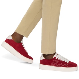 Model wearing Kurt Geiger London red suede trainers with white sole and embroidered script logo, styled with beige tailored trousers.