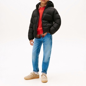 Model wearing Tommy Hilfiger black hooded puffer jacket, styled with red sweatshirt, light blue jeans and beige trainers.