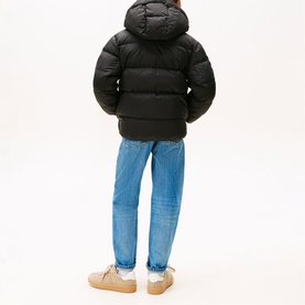 Model wearing Tommy Hilfiger black hooded puffer jacket, styled with blue jeans and beige trainers, seen from behind.