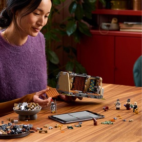 Model with Lego Icons set, assembling a detailed multi-coloured build at a wooden table with minifigures and accessories laid out nearby.