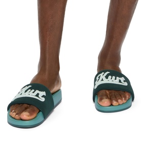 Model wearing Kurt Geiger London green slider sandals with white logo branding and teal ridged sole.