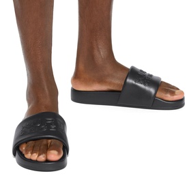 Model wearing Kurt Geiger London black leather slides with embossed logo strap and flat black sole