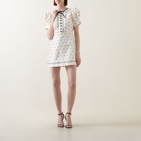 Model wearing Queens of Archive Suki Mini Dress in White Polka dot print with bow neckline, puff sleeves, and scallop trim; styled with black strappy