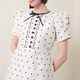 Model wearing Queens of Archive Suki Mini Dress in White Polka with black polka dots, bow appliqu&eacute; neckline, scallop trim, and puff sleeves.