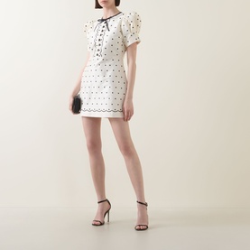 Model wearing Queens of Archive Suki Mini Dress in White Polka with bow neckline, styled with black heeled sandals and black clutch bag.