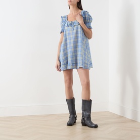 Model wearing Damson Madder Harlow Check Mini Dress in blue check with ruffle trim and short sleeves, styled with dark knee-high boots